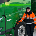 Rekka-auto edessä nainen pipo päässä ja katsoo kohti kameraa ja hymyilee.
