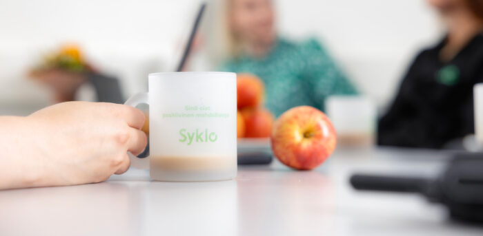 Lähikuva kädestä, joka pitelee pöydän päällä olevaa Syklon kahvimukia. Taustalla näkyy himmeänä omenoita sekä kaksi ihmistä.